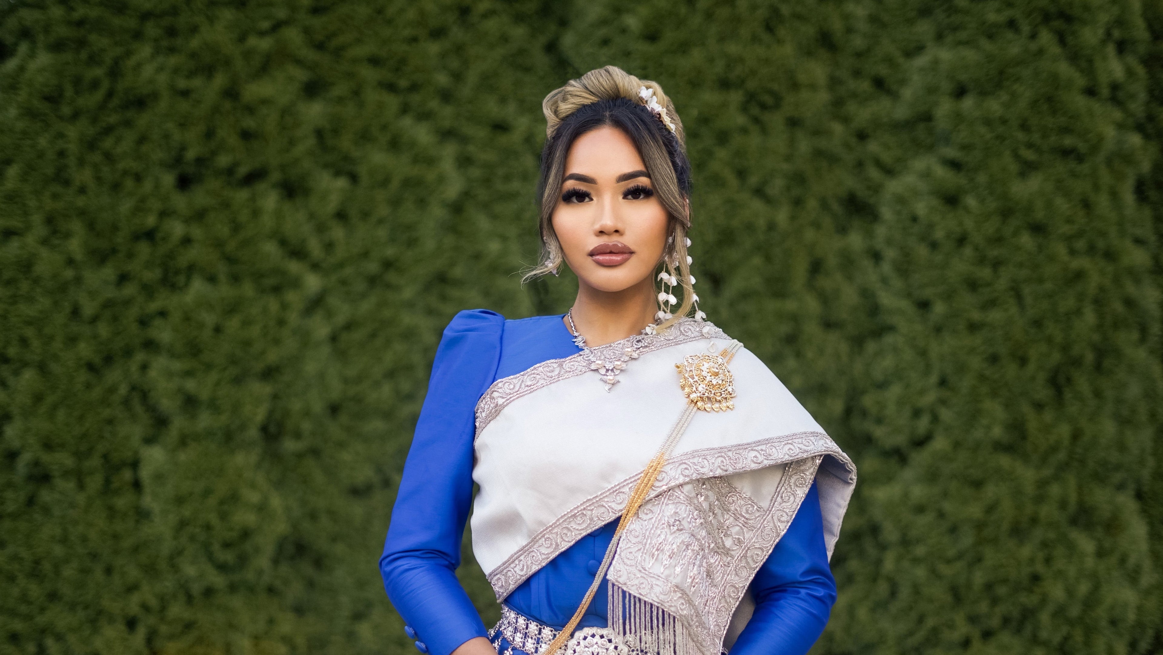 Woman in traditional blue and white outfit standing against a green hedge.